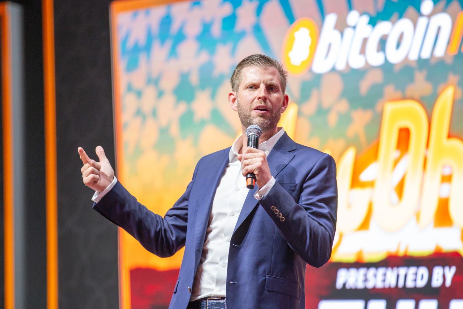 Eric Trump presenting at Bitcoin conference with Bitcoin symbol and financial charts representing institutional demand and supply scarcity.