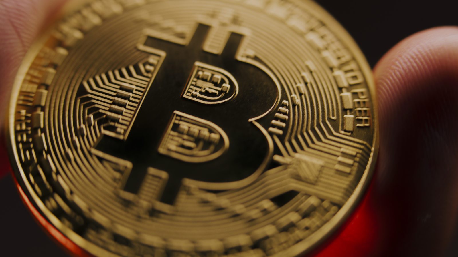 A dramatic close-up photo of a physical Bitcoin (BTC) coin being held between fingers. The image represents the cryptocurrency, which is the subject of an article about the acceleration of its adoption by sovereign nations in 2025.