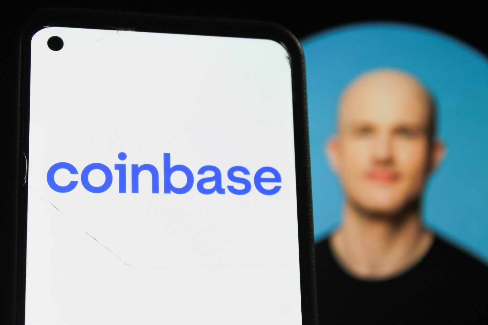 A smartphone in the foreground displays the blue Coinbase logo, with a blurred portrait of the company's CEO, Brian Armstrong, visible in the background. The image illustrates the connection between the company and its leader, whose "super app" strategy is the focus of the article.