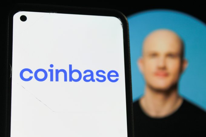 A smartphone in the foreground displays the blue Coinbase logo, with a blurred portrait of the company's CEO, Brian Armstrong, visible in the background. The image illustrates the connection between the company and its leader, whose "super app" strategy is the focus of the article.
