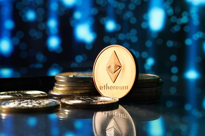 A physical representation of an Ethereum (ETH) coin is shown in focus, with other coins stuck around it on a reflective surface against a glowing blue digital background. The image represents the cryptocurrency that is the subject of an article about significant ETF outflows and a recent price dip.