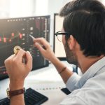 A trader, seen from behind, analyzes a bearish candlestick chart on a computer monitor while holding a physical Bitcoin. The image represents a crypto investor assessing the market's structural tailwinds, as discussed in the October 2025 article, following a major leverage crash.