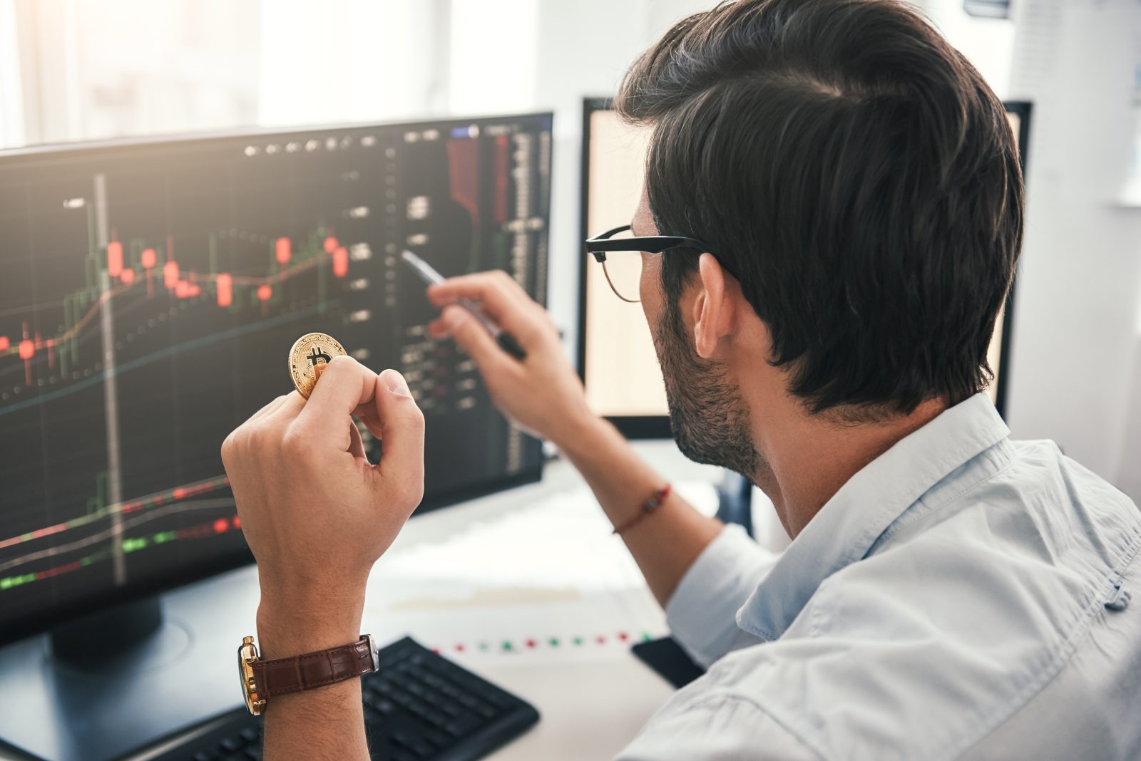 A trader, seen from behind, analyzes a bearish candlestick chart on a computer monitor while holding a physical Bitcoin. The image represents a crypto investor assessing the market's structural tailwinds, as discussed in the October 2025 article, following a major leverage crash.