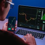 A crypto trader analyzes financial charts on both a laptop and a smartphone, where "BUY" and "SELL" buttons are visible. The image represents the active analysis of market data as the October 2025 article discusses the divergent performance and bullish sentiment for cryptocurrencies like XRP and Solana compared to Bitcoin and Ether.