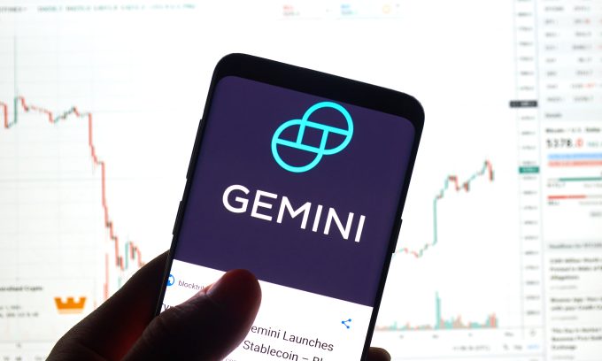 A smartphone displays the logo for the Gemini cryptocurrency exchange. In the background, a blurred financial chart is visible, representing the market analysis of crypto exchange stocks mentioned in the October 2025 article, which details Citi's price target cut for Gemini (GEMI) due to slowing volume.