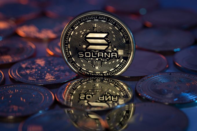 A close-up photograph of a physical Solana (SOL) coin standing upright among other coins on a reflective surface. The image represents the cryptocurrency that is the subject of a 2025 article about its potential to become a key network for Wall Street.