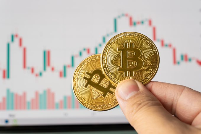A hand holds two physical Bitcoin coins in front of a blurred financial candlestick chart. The image represents the crypto market, which is the subject of an SKN market wrap (November 2025) detailing Robert Kiyosaki's $250K Bitcoin price target.