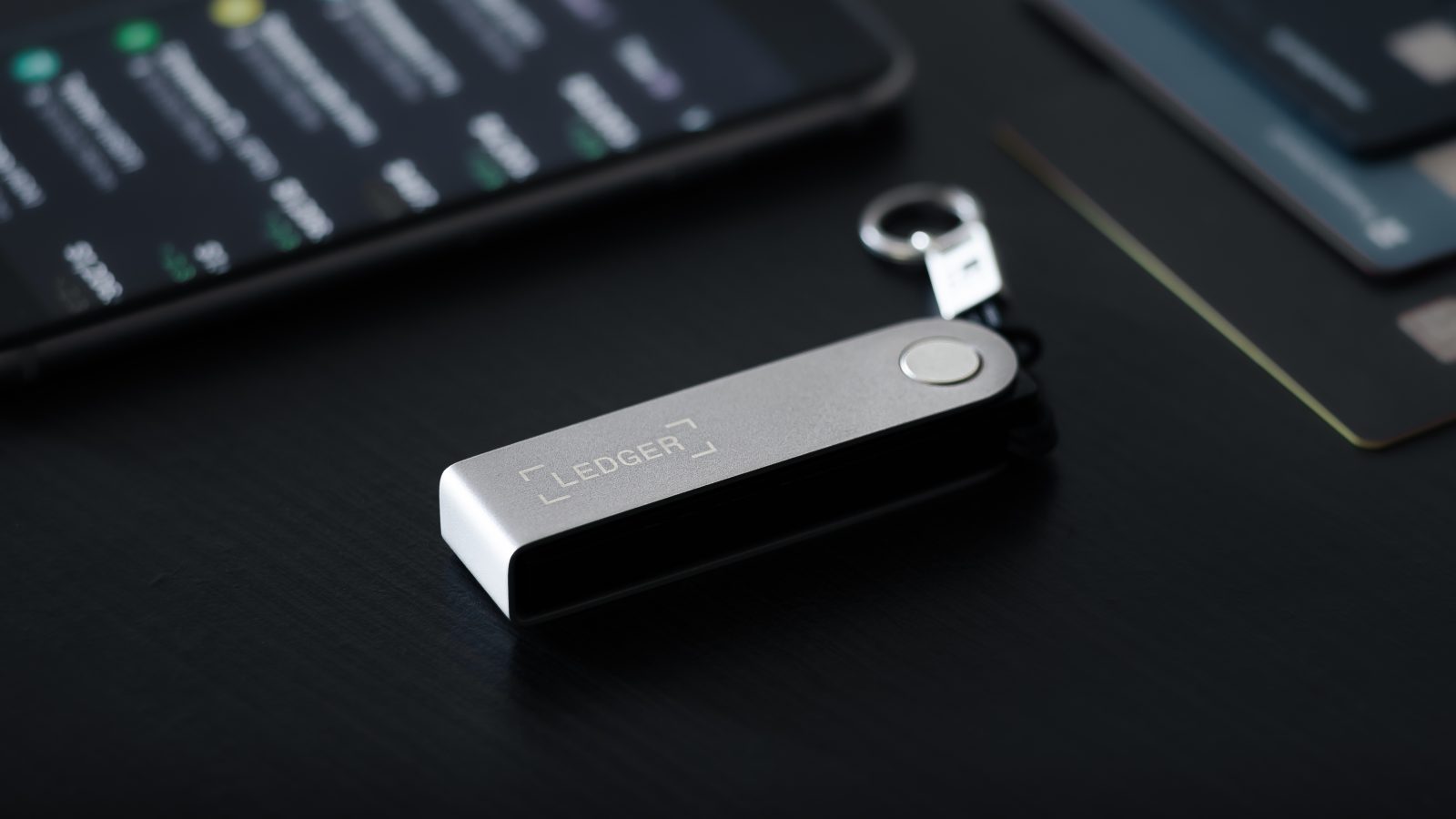 A Ledger hardware crypto wallet is shown on a desk next to a smartphone. The image represents the security product at the center of an SKN article from November 2025, detailing Ledger's plans for a New York listing (IPO) as record crypto hacks fuel demand for its devices.