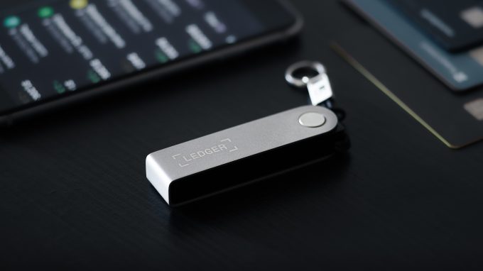 A Ledger hardware crypto wallet is shown on a desk next to a smartphone. The image represents the security product at the center of an SKN article from November 2025, detailing Ledger's plans for a New York listing (IPO) as record crypto hacks fuel demand for its devices.