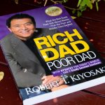The book "Rich Dad Poor Dad" by Robert Kiyosaki is shown. The image identifies the author, who, as detailed in the SKN article from November 2025, is targeting a $250K Bitcoin price and $27K gold price, calling them "real money" hedges against an impending market crash.