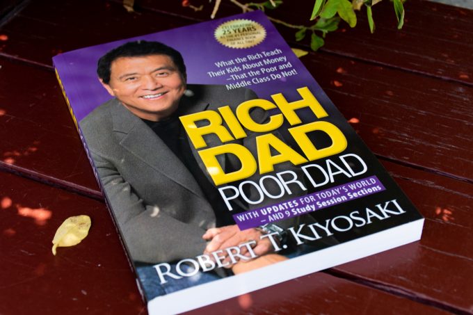 The book "Rich Dad Poor Dad" by Robert Kiyosaki is shown. The image identifies the author, who, as detailed in the SKN article from November 2025, is targeting a $250K Bitcoin price and $27K gold price, calling them "real money" hedges against an impending market crash.
