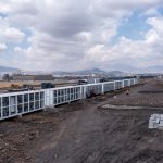 An expansive, outdoor view of a Bitcoin mining farm, showing long rows of white containerized mining units at a construction site.