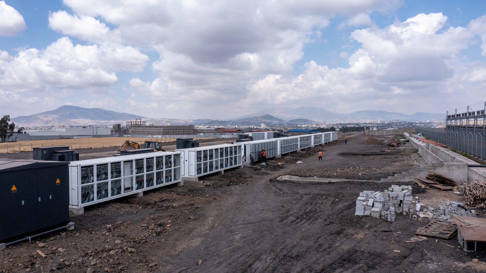 An expansive, outdoor view of a Bitcoin mining farm, showing long rows of white containerized mining units at a construction site.