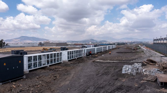 An expansive, outdoor view of a Bitcoin mining farm, showing long rows of white containerized mining units at a construction site.