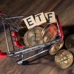 A miniature shopping cart filled with physical Bitcoin coins and wooden blocks spelling out 'ETF'.