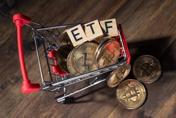 A miniature shopping cart filled with physical Bitcoin coins and wooden blocks spelling out 'ETF'.