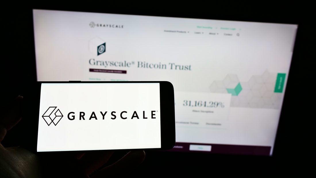 A smartphone screen displaying the Grayscale logo held in front of a blurred computer monitor showing the company's website.