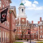 A sunny day view of the iconic brick buildings and clock tower of Harvard University's campus, with a Winthrop House flag flying.