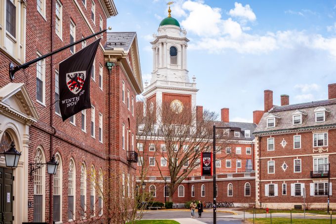 A sunny day view of the iconic brick buildings and clock tower of Harvard University's campus, with a Winthrop House flag flying.